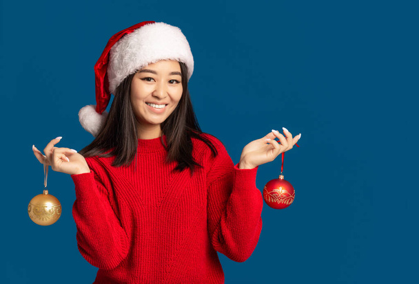 Onnellinen nainen pitää käsissään joulupalloja. Kaunis aasialainen nainen joulupukin hattu sinisellä taustalla, katsot kameraan ja hymyilet. Joulun käsite - Valokuva, kuva