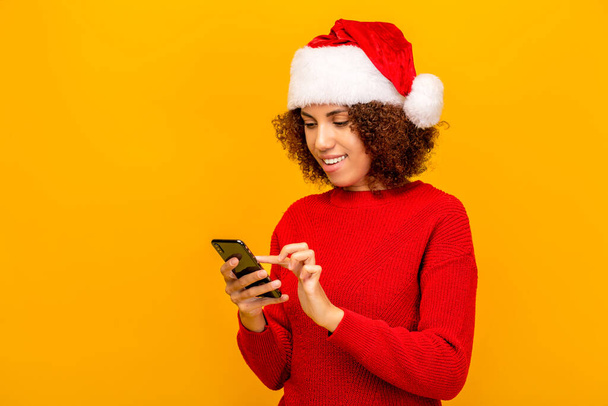 Young woman in santa claus hat holding mobile phone, reading holiday message, congratulates friends or relatives happy new year, standing on a orange background - Φωτογραφία, εικόνα