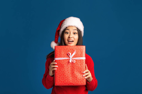 Surprised happy woman holds in hands christmas gift. Beautiful Asian woman in santa claus hat on blue background, Looking at the camera. Xmas concept - Фото, изображение
