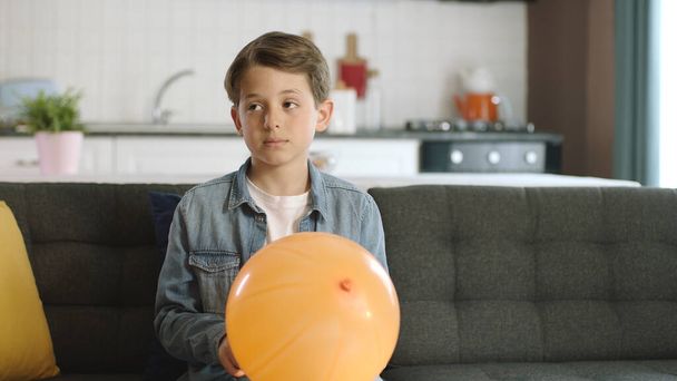 Auf einem grauen Sofa mit einem orangefarbenen Luftballon in der Hand, bereit für eine Geburtstagsfeier. Ein einsamer, freundlicher, verschlossener nerdy boy starrt auf die leere Werbefläche links vom Bildschirm. - Foto, Bild