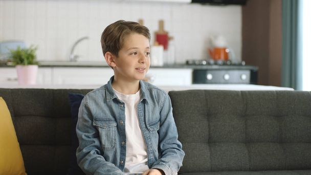 Portret szczęśliwego, pięknego uśmiechniętego dziecka. Piękny chłopiec siedzący samotnie na kanapie w domu patrzy na pustą przestrzeń reklamową po prawej stronie ekranu. - Zdjęcie, obraz