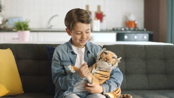 Kotona yksin, pieni poika ilman ystäviä leikkii lelu tiikerillä.Ruskea lelu tiikeri lapsen kädessä. Lapsi leikkii lelu tiikeri olohuoneessa. - Valokuva, kuva