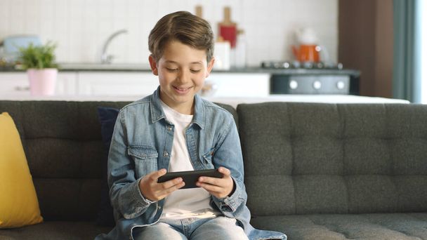 Boy using phone to play games. Little funny boy playing smartphone game while sitting on sofa at home, next generation games, happy childhood. - Foto, immagini