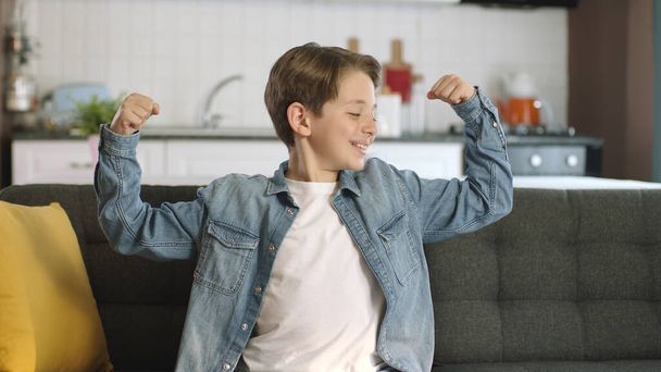 小さな男の子は、カメラに彼の腕の筋肉を示しています。家でカメラに足、筋肉、強さを示す小さな男の子の肖像画。. - 写真・画像