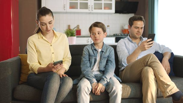 Exigiendo atención de sus padres jóvenes, el niño está aburrido con la difícil situación de sus padres adictos a la tecnología. Pareja adicta a la tecnología que mira sus cuentas de redes sociales en sus teléfonos inteligentes. - Foto, imagen