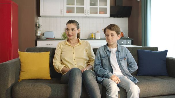 Young mother and little boy sitting on sofa with mother in living room of their home. He smiles and looks at the empty advertising space to the left of the screen. - Zdjęcie, obraz