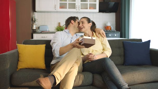 Woman surprises her young husband by giving a gift wrap while watching an exciting movie on TV.The man is happy to see the gift and kisses his young wife.Gift giving, mother's or father's day concept. - Foto, afbeelding
