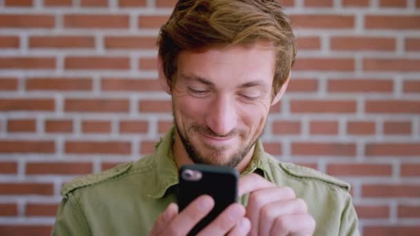 Man, smartphone and typing on mobile app with brick wall background in city, streaming social media or happy online communication. Young person, smile and website job search on phone in Los Angeles. - Footage, Video