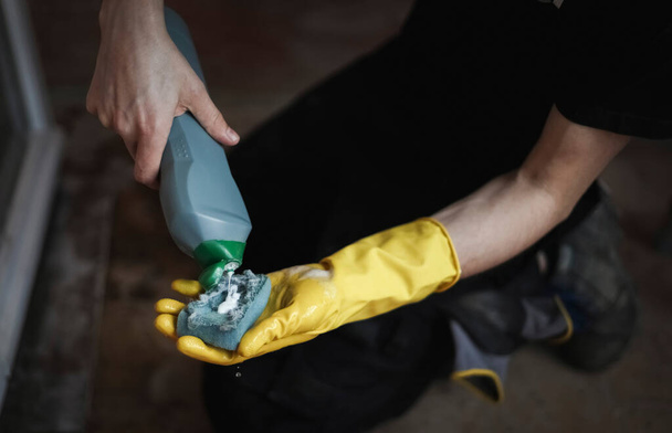 The hands of a caucasian young man in yellow gloves pour soapy liquid from a gray bottle onto a sponge while sitting on his knees on the floor, close-up side view with selective focus. The concept of home renovation, washing window frames. - Photo, image
