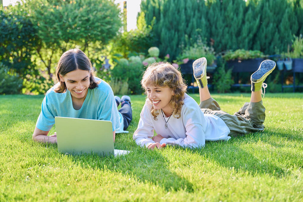 Two teenage friends of students lying on grass with laptop, in backyard, guy and girl 16, 17 years old study together. Friendship, youth, technology, high school, college, lifestyle concept - Фото, изображение