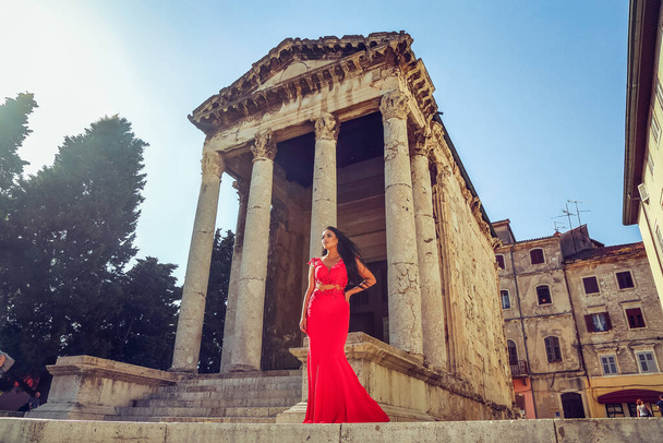 Beautiful young woman in a long red dress enjoys a walk in the city and poses next to an old monument - Fotoğraf, Görsel