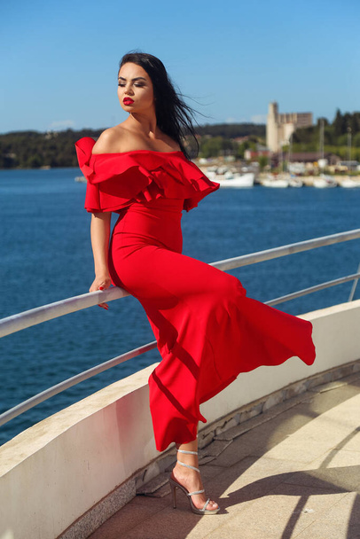 Beautiful young black-haired woman in a long red dress is enjoying herself on a yacht looking at the sea - Fotó, kép