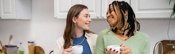 cheerful multiethnic lesbian couple holding cups of coffee and looking at each other in kitchen, banner - Fotografie, Obrázek