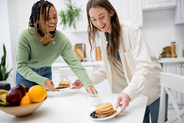 fröhliche lesbische Frau serviert Teller mit leckeren Pfannkuchen in der Nähe der afrikanisch-amerikanischen Freundin  - Foto, Bild