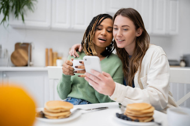 cheerful lesbian woman showing smartphone to surprised african american girlfriend during breakfast  - 写真・画像