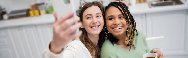 wesoła lesbijka robi selfie na smartfonie z radosną afrykańską dziewczyną, sztandar - Zdjęcie, obraz