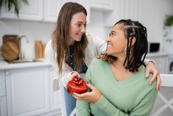boldog leszbikus nő kezében piros ajándék doboz közelében mosolygós afro-amerikai barátnő  - Fotó, kép