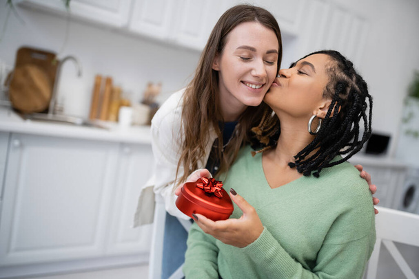Afrikaans amerikaans lesbisch vrouw zoenen wang van vrolijke vriendin en holding hartvormige gift box - Foto, afbeelding