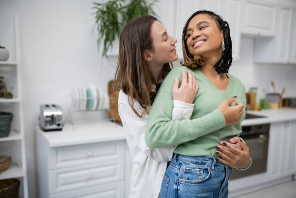 happy lesbian woman embracing smiling african american girlfriend in kitchen  - Photo, image