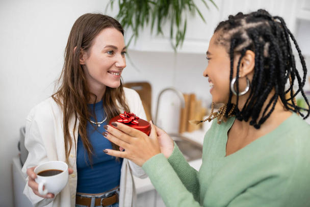 african american lesbian woman holding red gift box near happy girlfriend with cup of coffee on valentines day - Valokuva, kuva