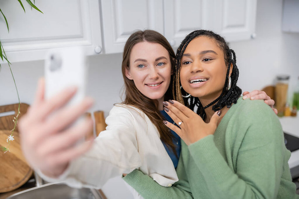 lesbiana mujer tomando selfie mientras alegre africana americana novia mostrando compromiso anillo en dedo  - Foto, imagen