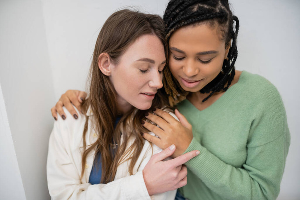 young interracial and lesbian women hugging each other near white wall - Fotografie, Obrázek