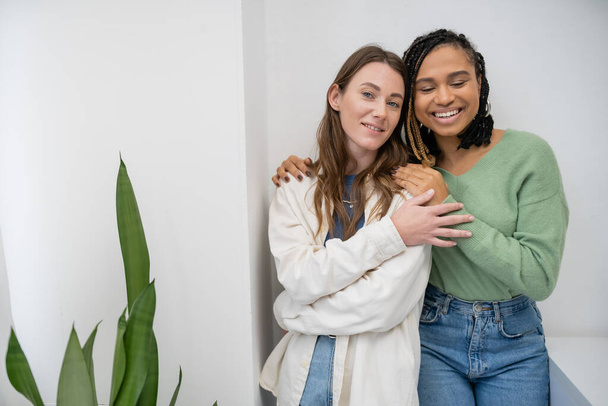 multiethnic and lesbian women smiling while hugging near green plant at home - Φωτογραφία, εικόνα