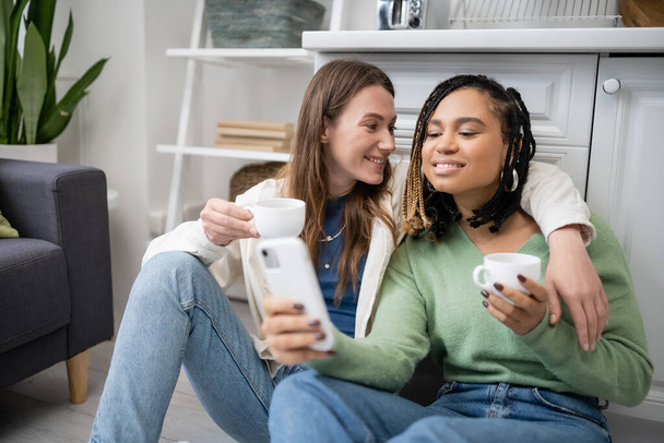 lesbisch afrikaans amerikaans vrouw holding smartphone in de buurt gelukkig vriendin met beker terwijl zitten op keukenvloer - Foto, afbeelding