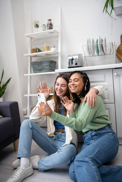 feliz interracial e lésbicas mulheres acenando as mãos durante chamada de vídeo enquanto sentado no chão da cozinha - Foto, Imagem