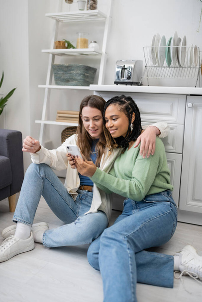 african american lesbian woman using smartphone near smiling girlfriend while sitting on kitchen floor - Фото, изображение