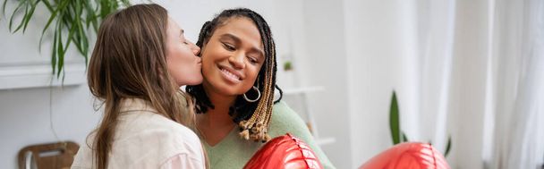 happy lesbian woman kissing cheek of pleased african american girlfriend near red balloons on valentines day, banner - Foto, Imagen