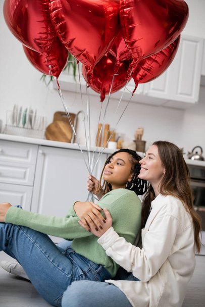 happy interracial and lesbian women sitting near red heart-shaped balloons on valentines day  - Photo, Image