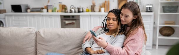młoda lesbijka wiadomości na smartfonie podczas odpoczynku na kanapie z afrykańską dziewczyną, baner  - Zdjęcie, obraz