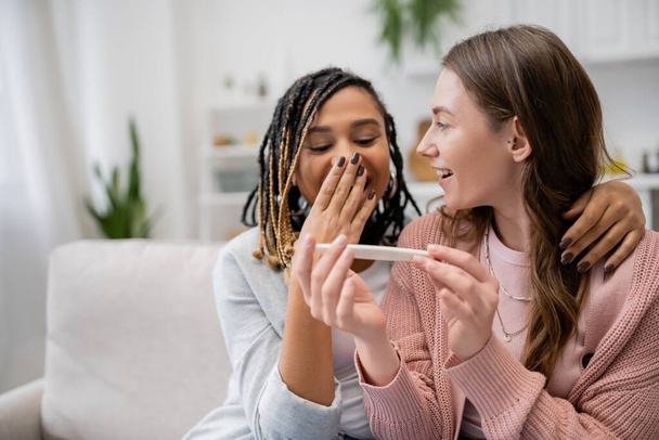 изумленная африканская американка и лесбиянка, прикрывающая рот, глядя на тест на беременность рядом с веселой девушкой   - Фото, изображение
