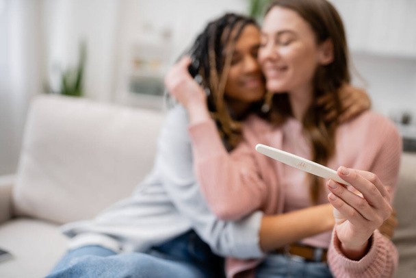 cheerful african american and lesbian woman hugging girlfriend with pregnancy test on blurred background  - Photo, Image