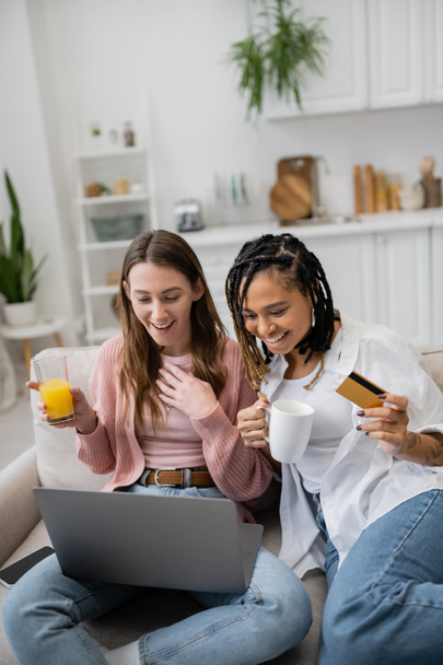 オンラインショッピング中にラップトップを使って幸せなガールフレンドの近くにクレジットカードを保持する若いアフリカ系アメリカ人レズビアン女性  - 写真・画像