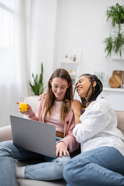 cheerful african american lesbian woman leaning on happy girlfriend using laptop while working from home  - Valokuva, kuva