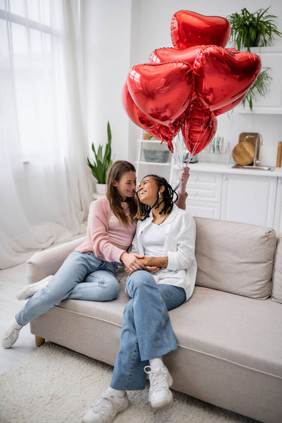 glückliche lesbische Frau hält herzförmige Luftballons und sitzt mit afrikanisch-amerikanischer Freundin auf der Couch  - Foto, Bild