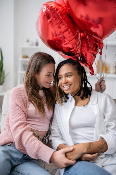 happy lesbian woman holding heart-shaped balloons and sitting on couch with african american girlfriend on valentines day  - Foto, Imagem