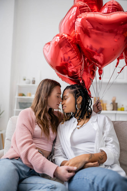 sensual lesbian woman holding heart-shaped balloons and sitting on couch with african american girlfriend on valentines day  - Fotografie, Obrázek