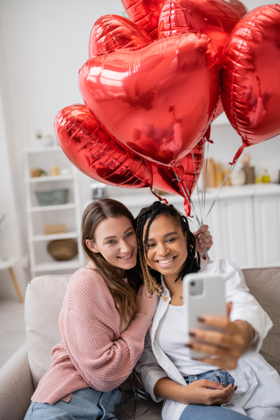 positive and multiethnic lesbian couple taking selfie near red balloons on valentines day  - Photo, Image