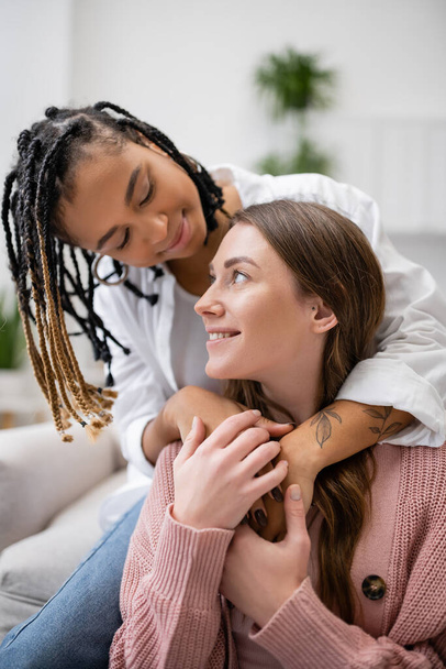 getatoeëerd Afrikaans amerikaans lesbisch vrouw met dreadlocks knuffelen gelukkig vriendin in woonkamer  - Foto, afbeelding