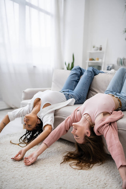 Positives und multiethnisches lesbisches Paar liegt kopfüber auf Couch im Wohnzimmer - Foto, Bild