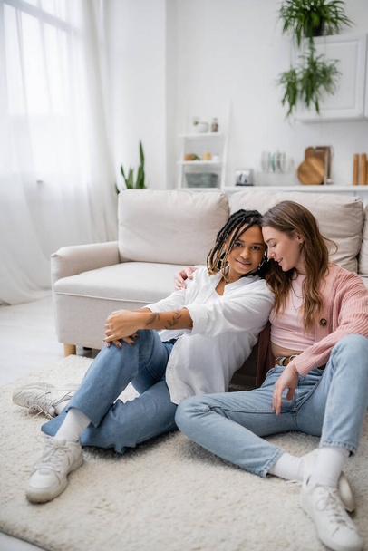 young lesbian woman sitting with tattooed african american girlfriend on carpet near couch  - Photo, Image