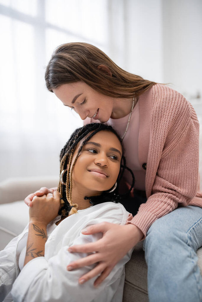 smiling lesbian woman sitting on couch and hugging tattooed african american girlfriend - Foto, afbeelding