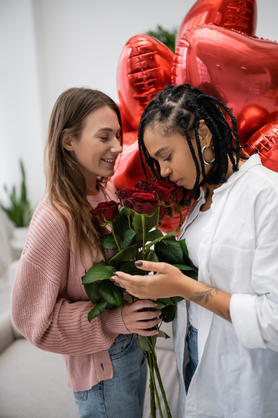 felice donna lesbica che tiene rose rosse vicino tatuato fidanzata africana americana e palloncini il giorno di San Valentino - Foto, immagini