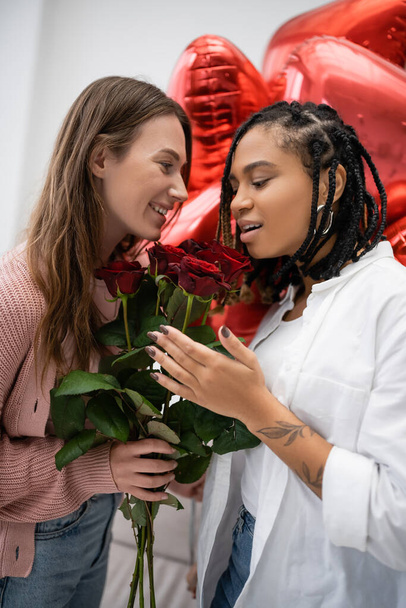 sonriente lesbiana mujer sosteniendo rosas rojas cerca asombrado africano americano novia y globos en valentines día - Foto, Imagen