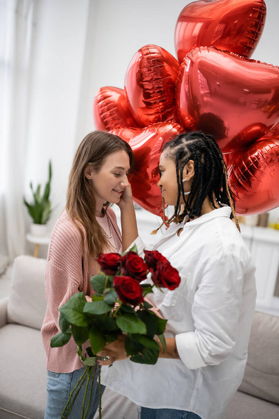 donna afroamericana lesbica toccando guancia di fidanzata con rose rosse in piedi vicino palloncini il giorno di San Valentino - Foto, immagini