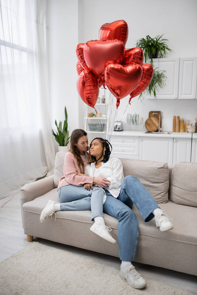 multiethnic lesbian women sitting on couch near heart-shaped balloons on valentines day  - Фото, зображення