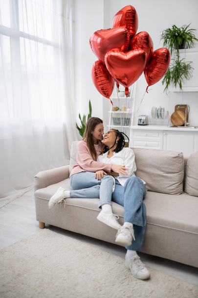 happy multiethnic lesbian women sitting on couch near heart-shaped balloons on valentines day  - Foto, Bild